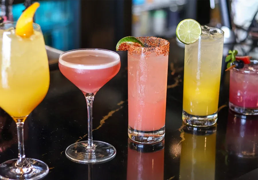 Five vibrantly colored, garnished cocktails on a dark reflective surface.