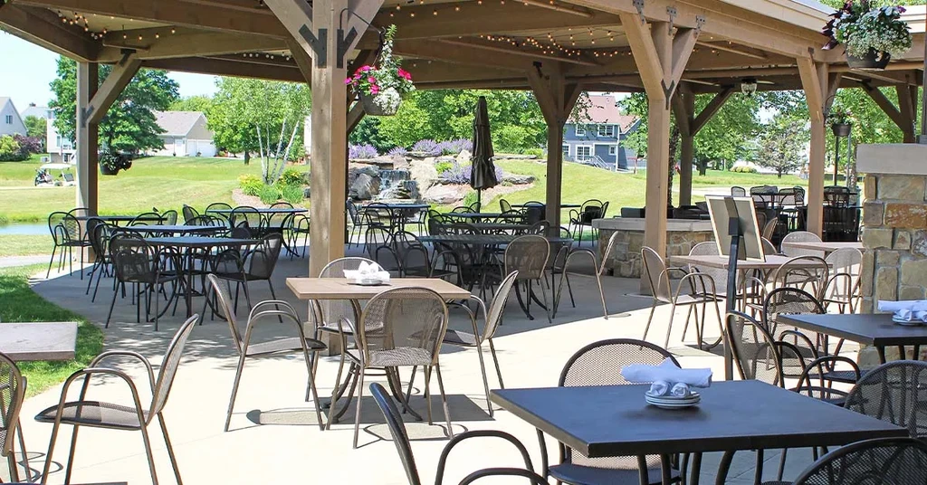 Sunny outdoor dining patio with pergola, pond, waterfall, and houses.