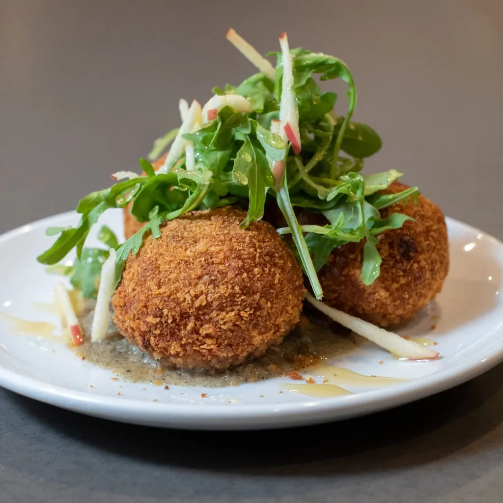 Three golden arancini topped with arugula, apple, and dark sauce.