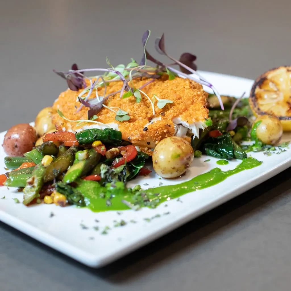 Artfully plated breaded main course with vegetables, potatoes, green sauce, lemon.