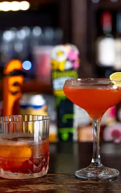 Two cocktails: dark amber in tumbler, vibrant orange in coupe.