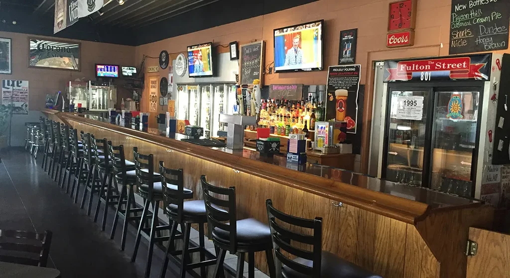 Empty bar interior with a long wooden counter, stools, liquor, and TVs.
