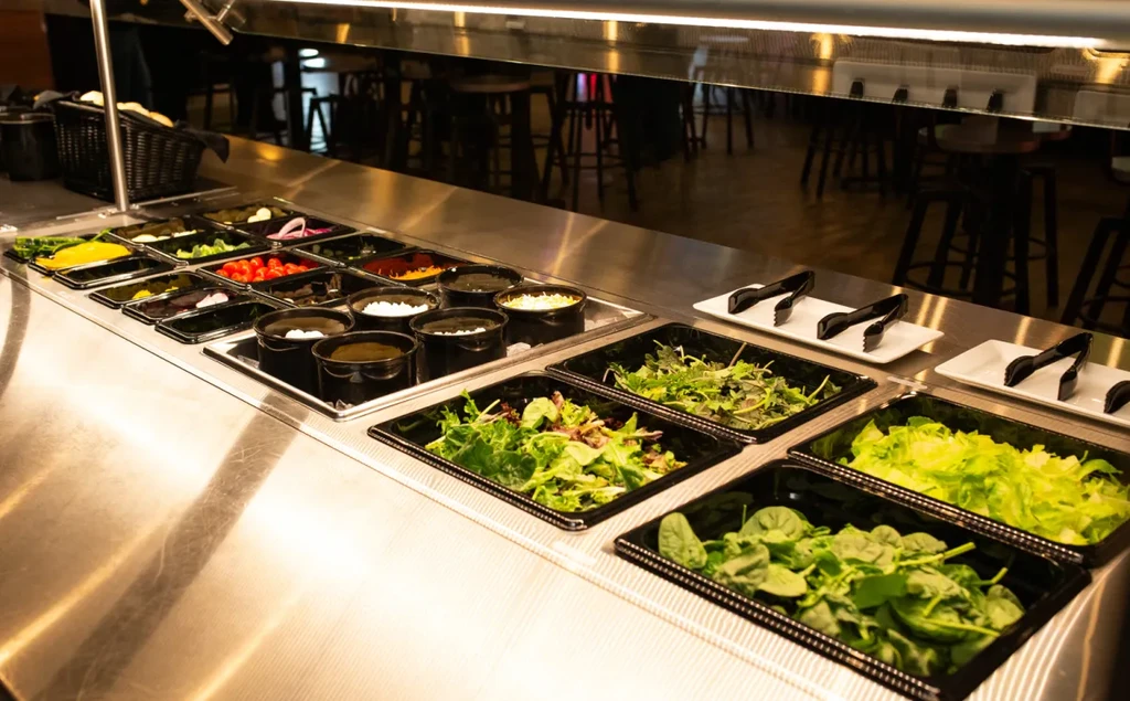 Well-lit, self-service stainless steel salad bar with fresh greens and toppings.