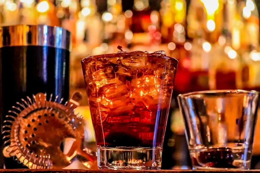 Dark amber cocktail in rocks glass with ice, shaker, blurred bar.
