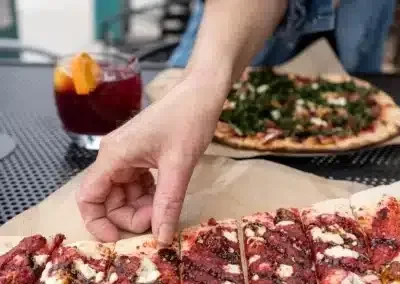 Hand picks slice from rectangular pizza; round pizza and red drink nearby.