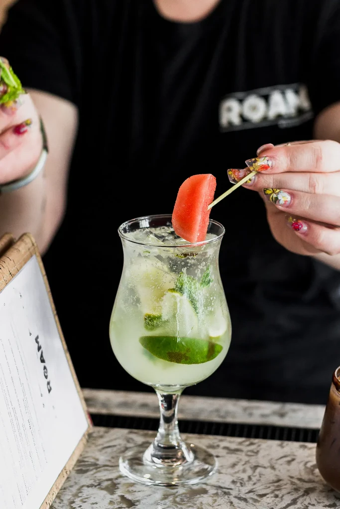 Person's hand garnishes clear cocktail with watermelon, lime, and mint.