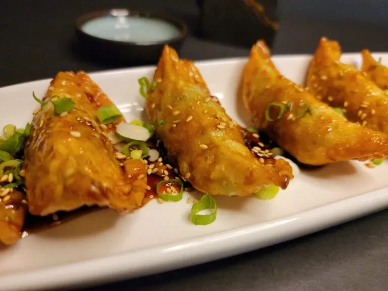 Golden-brown pan-fried dumplings, sauce, green onions, sesame seeds on white plate.