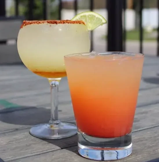 Two cold, garnished drinks on a rustic outdoor table in sunlight.