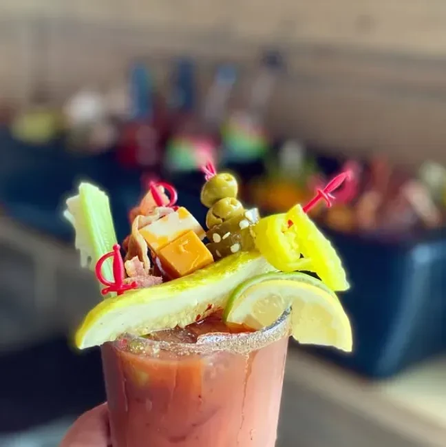 Hand holds a Bloody Mary cocktail garnished with celery, olives, cheese.