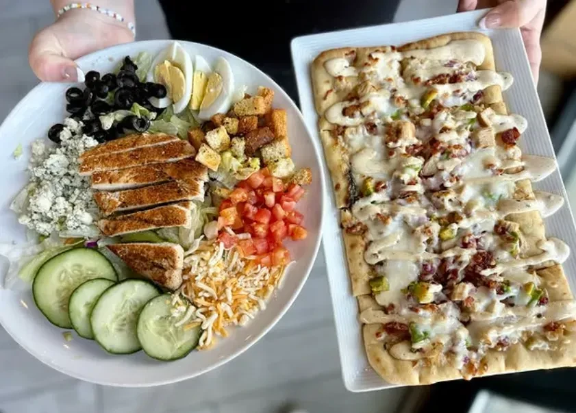 Two hands hold plates: one a chicken salad, the other a flatbread.