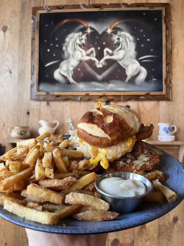 Hearty pretzel bun sandwich, fries, and a unicorn painting in background.