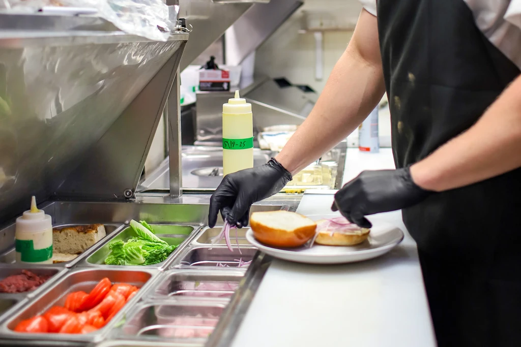 Gloved hands placing sliced red onions on a bun in a kitchen.
