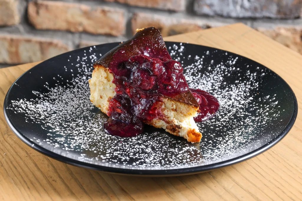 Slice of cheesecake with dark berry compote on a sugar-dusted plate.