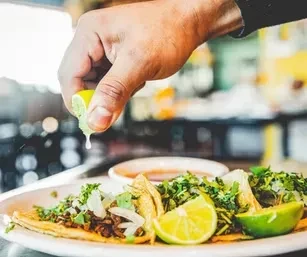 Hand squeezing lime over three tacos with meat, cilantro, and onions.