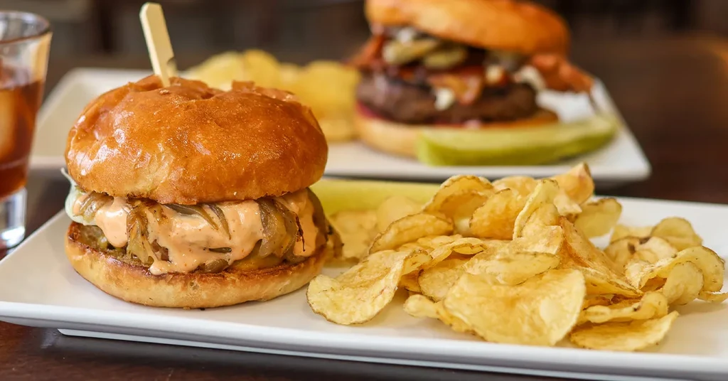 Close-up: Two gourmet burgers, chips, and a drink on a wooden table.