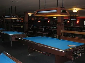 Dimly lit pool hall with multiple blue-felt tables under bright lights.