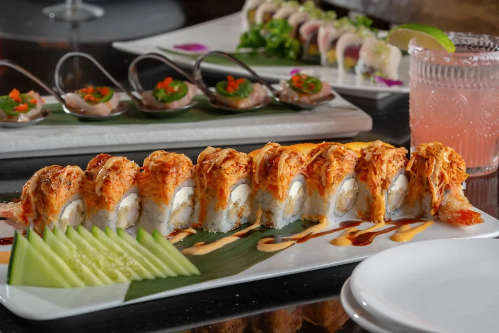 Orange-topped sushi, sashimi spoons, and a pink drink on a dark table.