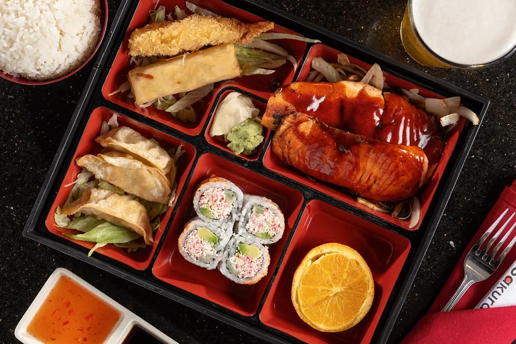 A black bento box meal containing various Asian dishes and beer.