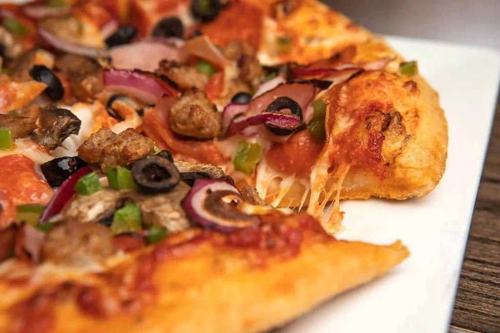 Close-up of loaded supreme pizza with cheese pull on white plate.