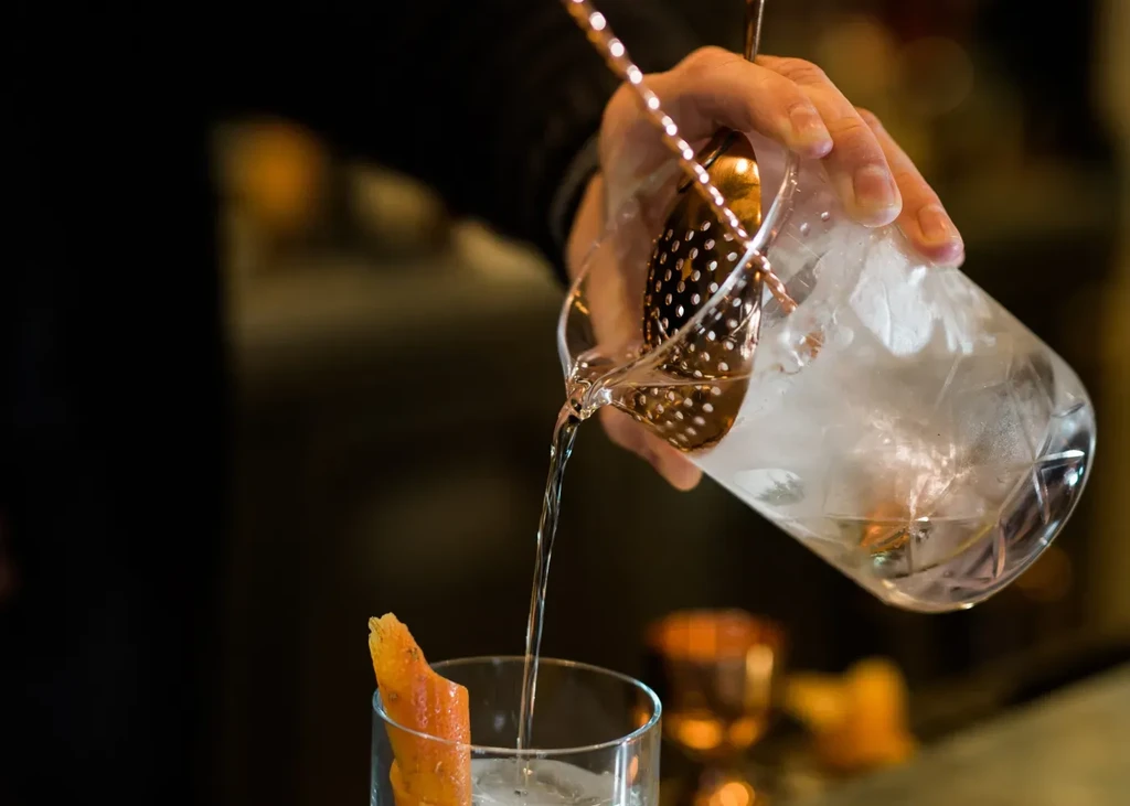 Close-up: Bartender's hands straining an iced drink with orange peel.