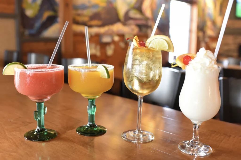 Four colorful cocktails in varied glasses on a wooden surface.