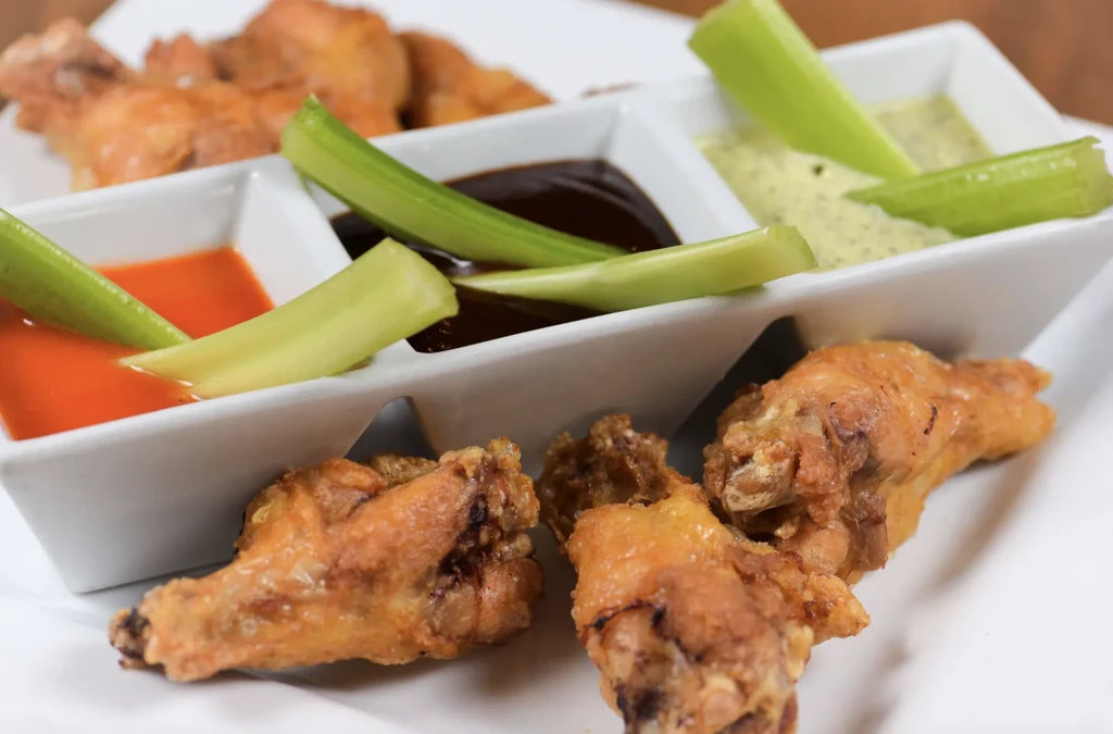 Close-up of golden-brown fried chicken wings, three dipping sauces, and celery.