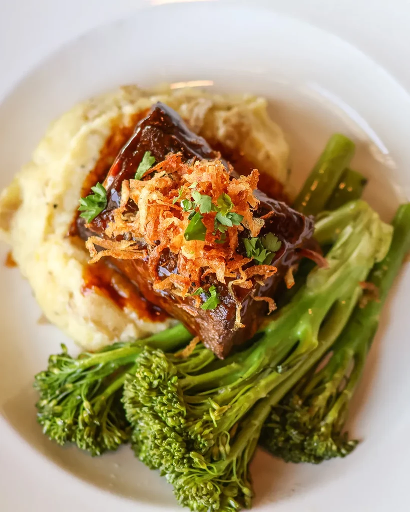Plated braised short rib, mashed potatoes, broccolini, and fried shallots.