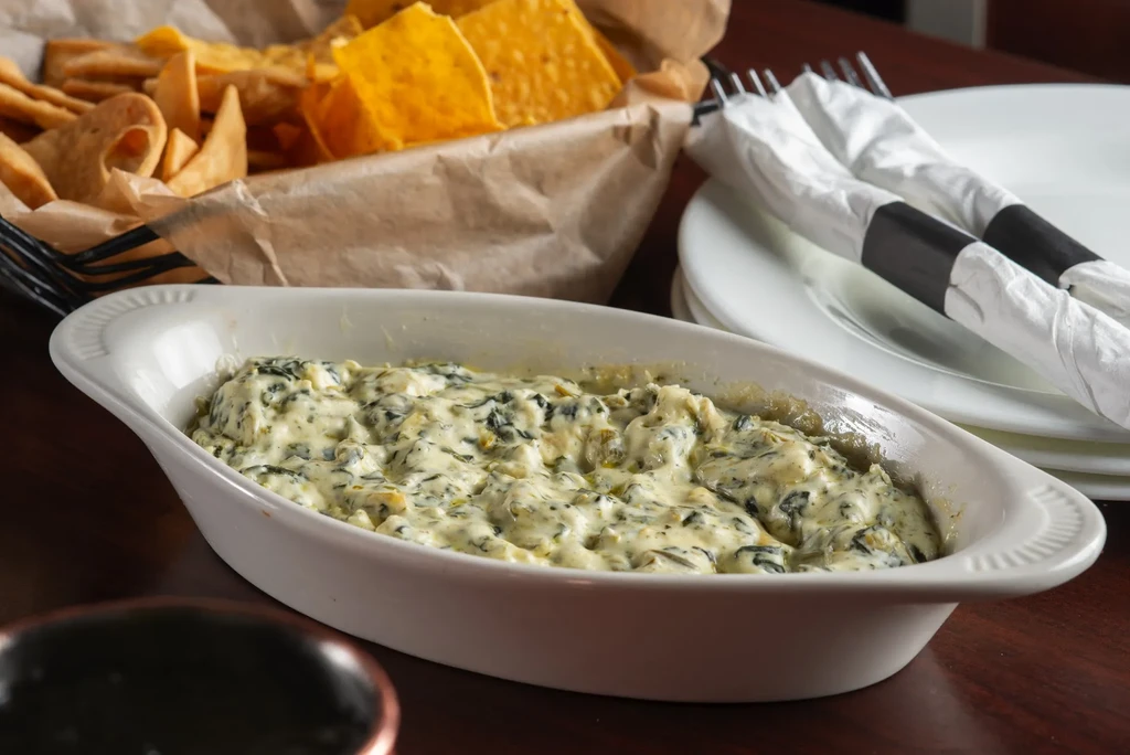 Spinach artichoke dip with chips, plates, and cutlery on a wooden table.