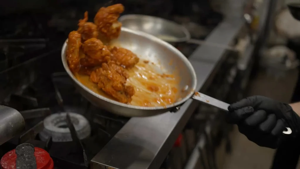 Gloved hand tosses fried chicken in orange-red sauce in a sauté pan.