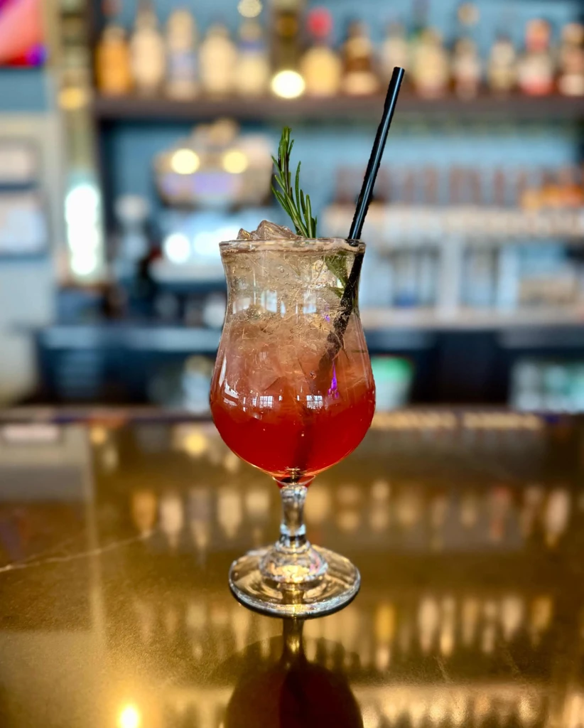 Red layered cocktail with rosemary and straw on bar counter.