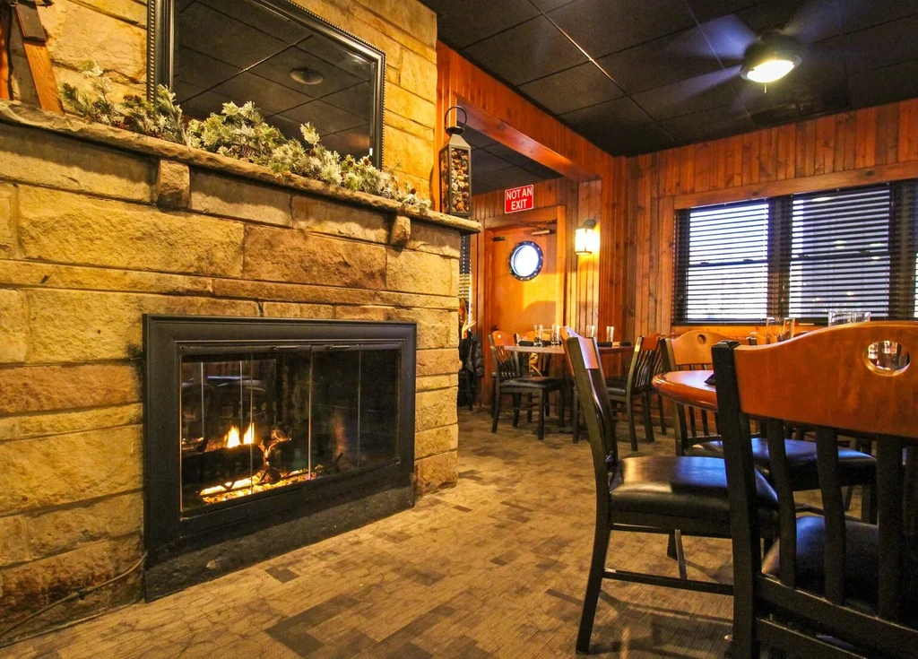 Stone fireplace burning in a rustic restaurant with wood walls and tables.