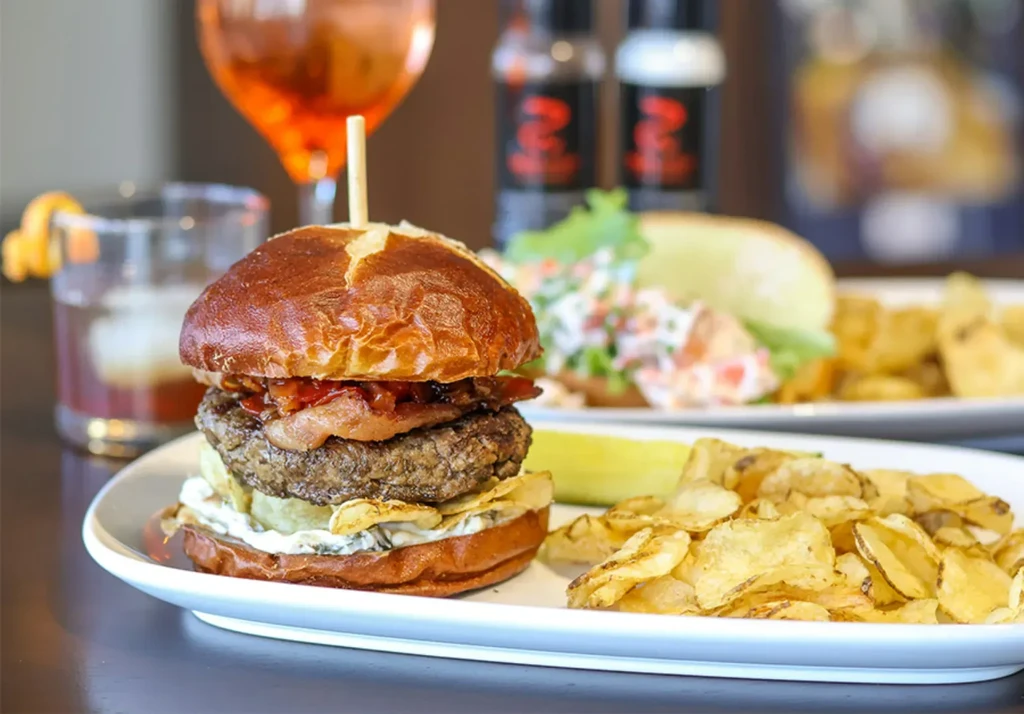 Gourmet pretzel bun burger, chips, pickle; blurred drinks in background.