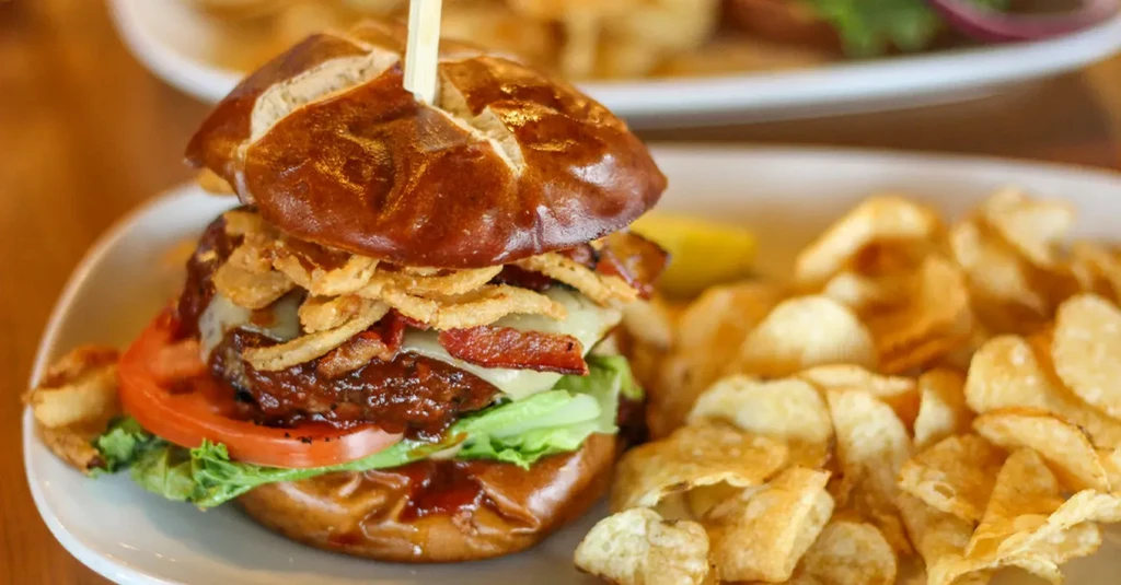 Close-up gourmet burger, pretzel bun, bacon, cheese, fried onions, potato chips.