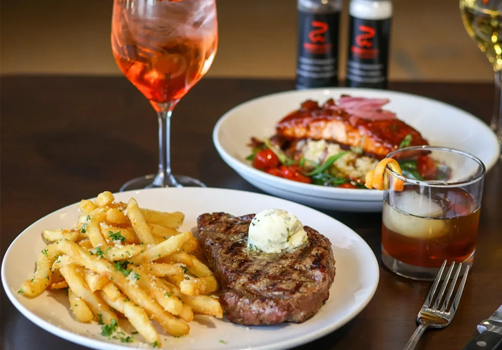 Grilled steak, fries, cocktails, and glazed salmon on a dark table.