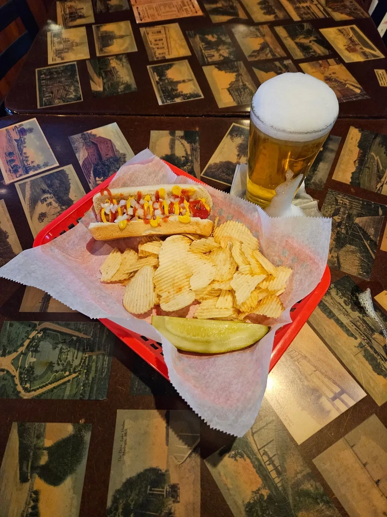 Hot dog, chips, pickle, and beer on a sepia image table.