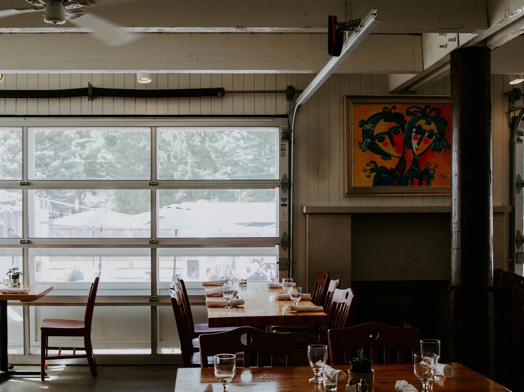 Empty restaurant interior with set wooden tables, natural light, and a colorful painting.