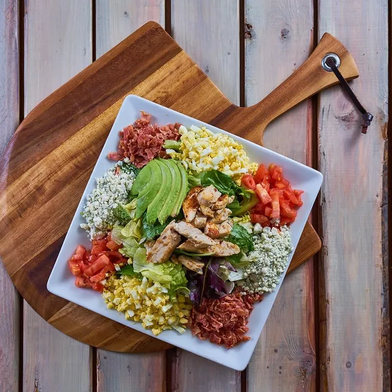 Elaborate Cobb salad with avocado, chicken, egg, bacon on rustic wood.