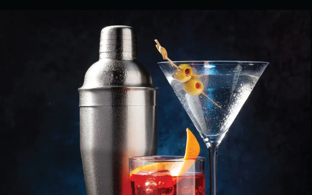 Still life of a cocktail shaker, martini, and orange-garnished drink.