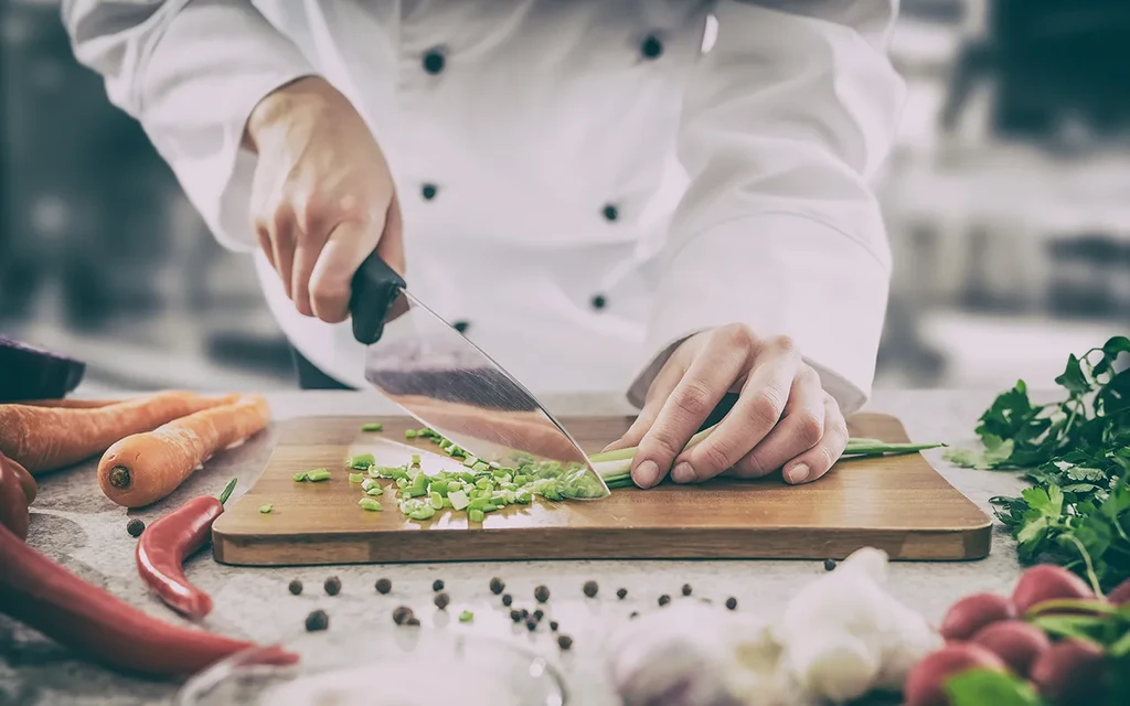 Chef's hands chopping green onions on a cutting board, surrounded by fresh vegetables.