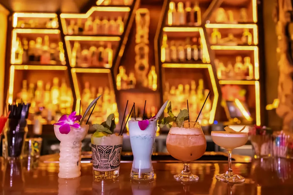 Five elaborately garnished cocktails on a reflective bar, against warm shelves.