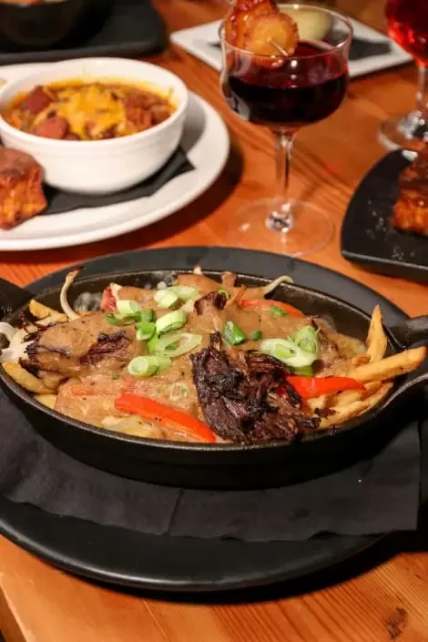 Cast-iron skillet of loaded fries, chili, and beverage on wood.