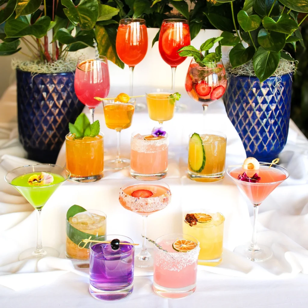 Thirteen colorful, garnished cocktails in various glasses on a tablecloth.