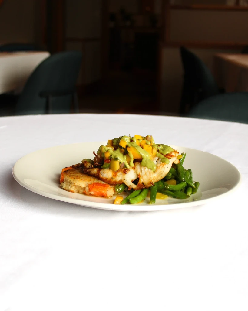 Plated white fish with mango salsa, green beans, and seafood patty.