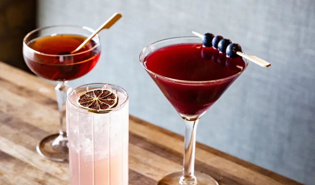 Three cocktails in coupe, highball, and martini glasses on wood.