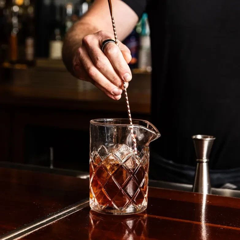 Bartender stirs an amber cocktail with ice in a patterned mixing glass.