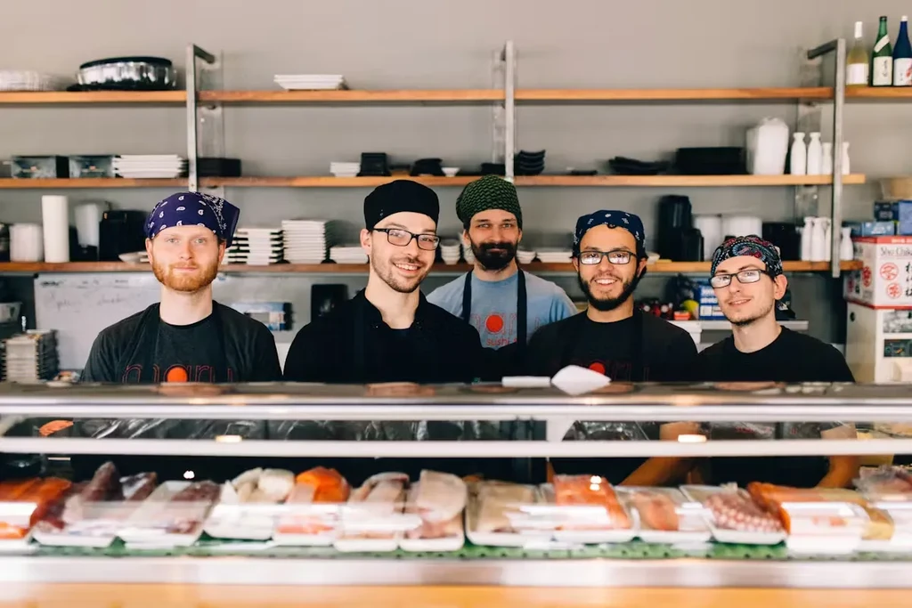 Five smiling chefs behind a sushi counter with raw fish display.
