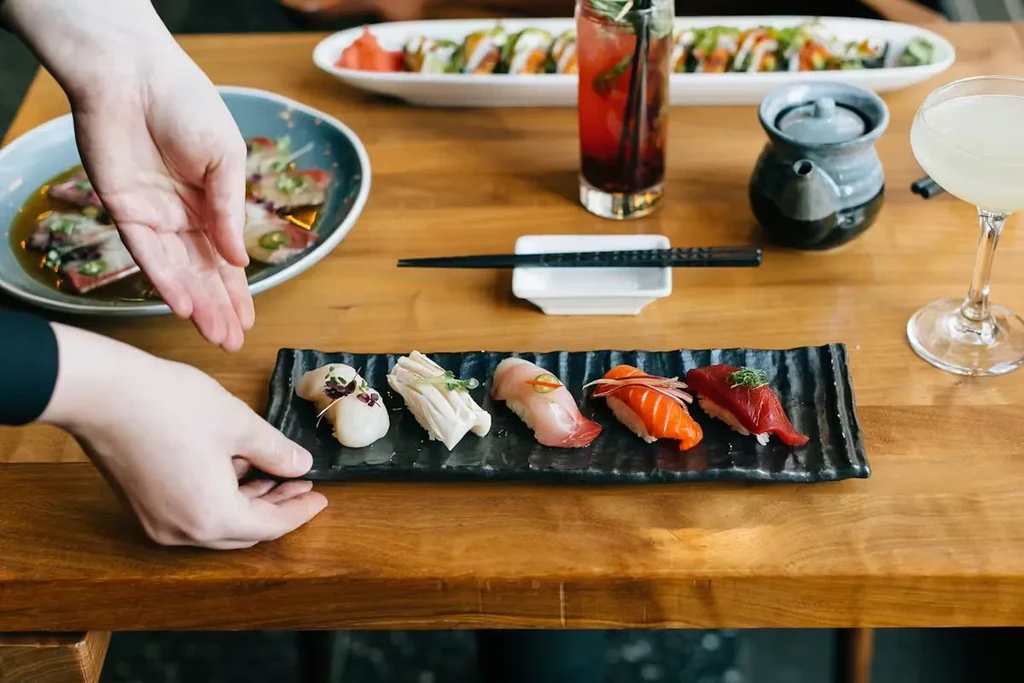 Hands placing nigiri sushi among Japanese dishes and drinks on a table.