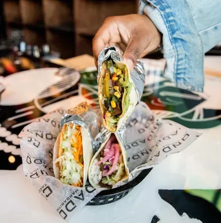 Hand holding one of three vibrant tacos in a black basket.