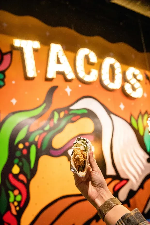 Hand holding a crispy taco; colorful mural and "TACOS" sign in background.
