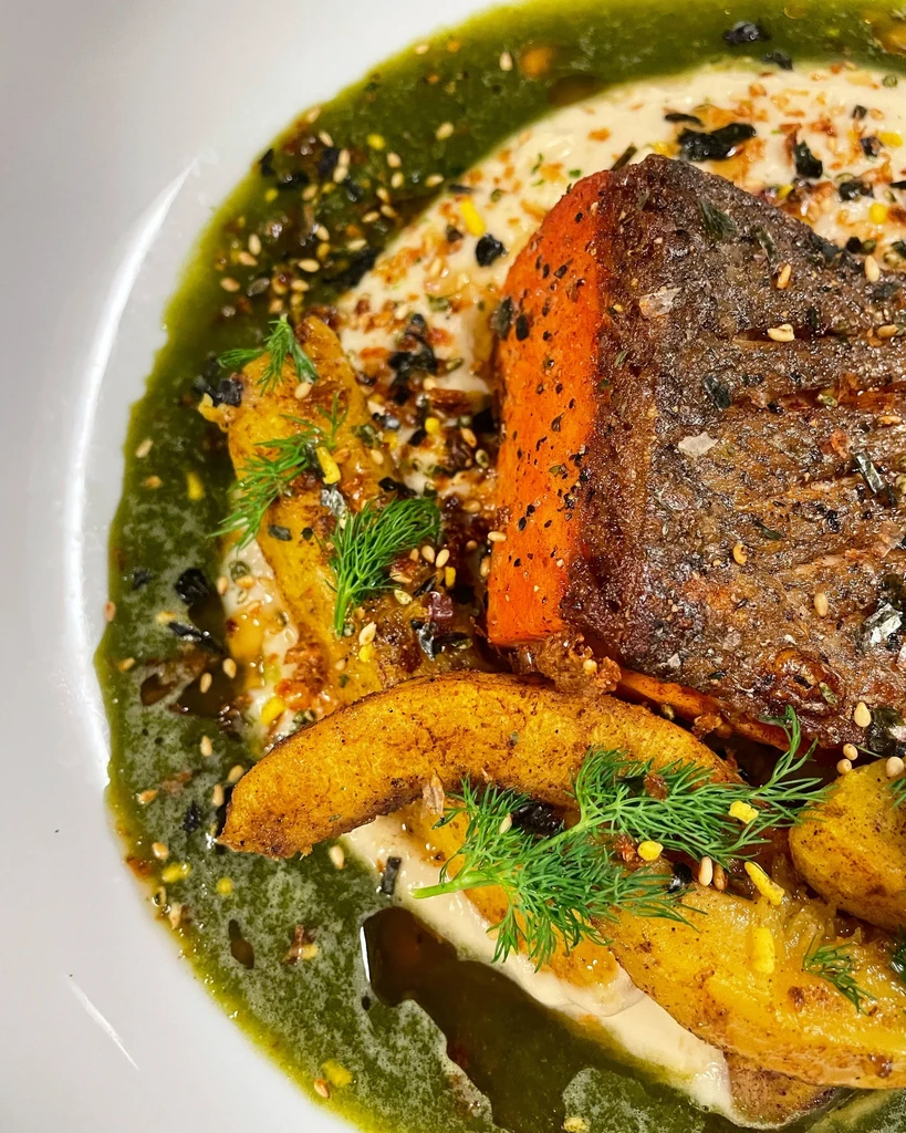 Close-up of seared fish, creamy base, roasted potatoes, dill, green sauce.